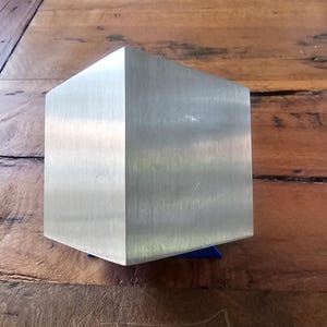 Pode incluir: Um cubo de prata com acabamento escovado, sentado em uma superfície de madeira.
