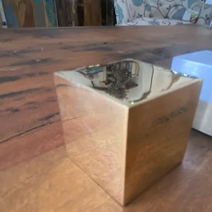 Può includere: Un cubo dorato lucido con una superficie riflettente è posto su un tavolo di legno. La superficie del cubo riflette l'ambiente circostante. Un altro cubo bianco è visibile sullo sfondo. I cubi sono oggetti decorativi.