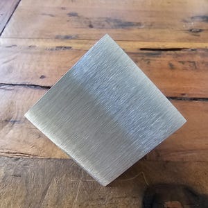 Pode incluir: Uma placa de metal prateado escovado com forma retangular. A placa está sobre uma superfície de madeira.