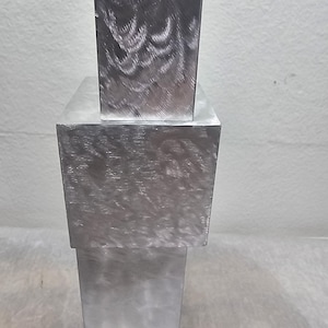 Pode incluir: Uma pilha de três blocos retangulares de metal escovado. O bloco superior é menor que o bloco do meio, e o bloco inferior é o maior. O metal tem um padrão texturizado e rodopiante. Os blocos são de cor prateada.