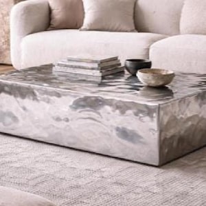 May include: A rectangular coffee table with a polished silver-toned metallic finish and a rippled, hammered surface texture. The table features a low-profile, block-like design with sharp edges and a reflective, undulating top surface.