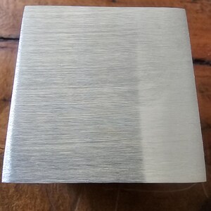 Pode incluir: Um objeto metálico retangular com acabamento escovado. O objeto tem uma cor prateada clara e uma forma ligeiramente trapezoidal. A superfície mostra linhas de grão horizontais.