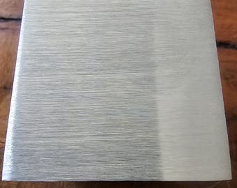 Könnte beinhalten: Ein rechteckiger Metallgegenstand mit gebürsteter Oberfläche. Das Objekt hat eine hellsilberne Farbe und eine leicht trapezförmige Form. Die Oberfläche zeigt horizontale Maserungslinien.