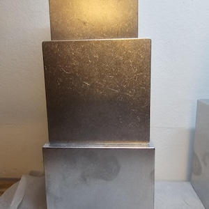 Pode incluir: Três cubos metálicos prateados empilhados verticalmente. O cubo maior está na base, os outros estão sobrepostos. As superfícies apresentam variações de textura e tom, refletindo a luz ambiente.
