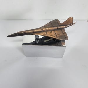 Puede incluir: Un modelo de avión de color bronce con un diseño elegante, montado sobre una base rectangular plateada. El avión presenta líneas de panel detalladas y una nariz puntiaguda. El modelo es un artículo decorativo.