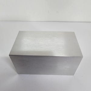 May include: A rectangular block made of brushed metal. The surface has a linear grain pattern, and the edges are sharp. The block is a light silver colour and sits on a white surface.