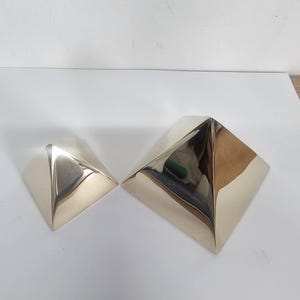 Pode incluir: Dois objetos polidos em forma de pirâmide de cor prateada. Os objetos são reflexivos, com uma superfície lisa. As pirâmides são colocadas sobre uma superfície branca.