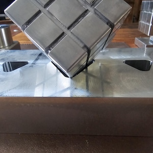 Puede incluir: Un cubo metálico con un patrón de rejilla, apoyado sobre una superficie metálica. El cubo está compuesto por bloques cuadrados más pequeños. La imagen muestra el diseño geométrico y la calidad reflectante del metal.