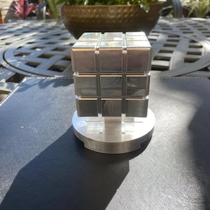 Pode incluir: Um cubo de quebra-cabeça prateado com um design de grade 3x3, montado em uma base circular. O quebra-cabeça é feito de metal e reflete a luz. A imagem é tirada ao ar livre, com um fundo desfocado de uma mesa e flores em vasos.