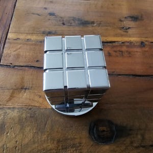 Puede incluir: Un cubo de metal plateado con un patrón de cuadrícula en cada lado. El cubo está sentado sobre una base redonda.