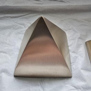 Pode incluir: Objeto metálico em forma de pirâmide com base plana e topo pontiagudo. A pirâmide tem uma cor bronze com um efeito gradiente, passando de um tom mais claro no topo para um tom mais escuro na base. Fundo branco texturizado.