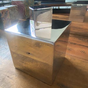 Pode incluir: Um cubo grande e brilhante, de cor prateada, com um cubo menor em cima. A superfície refletora dos cubos cria um efeito de espelho, refletindo o ambiente circundante. Os cubos estão colocados numa superfície de madeira.