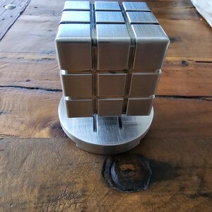 Pode incluir: Um quebra-cabeça de cubo metálico prateado, composto por pequenos blocos quadrados, apoiado sobre uma base circular. O objeto é exibido em uma superfície de madeira.