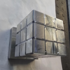 Könnte beinhalten: Eine metallische Würfelskulptur, die aus mehreren kleinen, gebürsteten Metallquadraten besteht. Die Quadrate sind so angeordnet, dass sie einen größeren Würfel bilden, der auf einer rechteckigen Basis montiert ist. Die Skulptur hat eine moderne, industrielle Ästhetik.