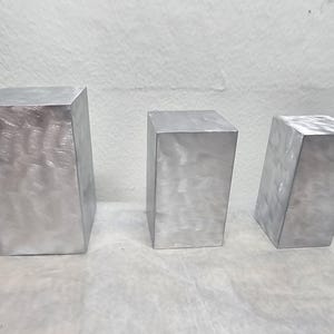 May include: Three silver-colored, rectangular blocks of varying sizes are arranged on a white surface. The blocks have a brushed metal appearance, with visible grain. The blocks are solid and have a simple, geometric design.