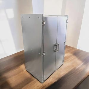 May include: A rectangular, brushed stainless steel cabinet featuring two vertical doors with a central metal handle. The structure is assembled with visible rivets along the side panel and includes three metal hinges on the right door frame.