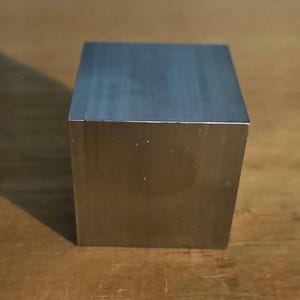 Peut inclure: Un cube métallique massif avec une finition brossée. Le cube est posé sur une surface en bois, avec une ombre projetée sur la gauche. Les bords du cube sont nets et bien définis, reflétant la lumière ambiante.