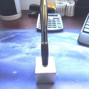 Pode incluir: Uma caneta prateada e preta com detalhes dourados está em uma base quadrada branca. A caneta tem uma pega escura e uma ponta dourada. O fundo inclui materiais de escritório, como uma calculadora e um grampeador.