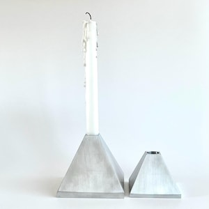 Pode incluir: Dois castiçais de metal na cor prateada com formato de pirâmide. O suporte mais alto contém uma vela cônica branca com gotas de cera visíveis na lateral. Ambos os suportes possuem um acabamento metálico escovado e uma base quadrada.