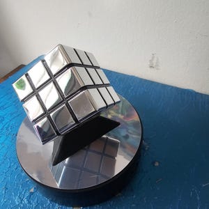 Pode incluir: Um cubo de Rubik prateado e refletivo está sobre uma base circular espelhada. Os quadrados do cubo refletem o ambiente. A base é suportada por um suporte triangular preto, tudo sobre uma superfície azul.