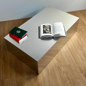 May include: A contemporary, mirrored coffee table. Two books are stacked on one end, a green book on top of a red one. An open book lies on the table's surface. The table is rectangular and angled, with a polished finish.