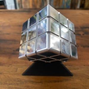 May include: A silver metal puzzle cube with a black base. The cube is made up of smaller squares that can be rotated and manipulated.