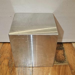 Pode incluir: Um cubo de metal prateado com acabamento escovado. O cubo está sobre uma superfície de madeira.