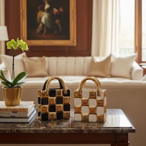 Könnte beinhalten: Zwei dekorative Keramikhandtaschen mit goldenen Griffen. Eine ist schwarz-gold kariert, die andere weiß-gold kariert. Sie stehen auf einem Marmortisch, mit einer goldenen Orchidee und Büchern links. Im Hintergrund sind ein Sofa und ein Gemälde zu sehen.
