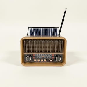 Può includere: Una radio in stile vintage con un involucro color oro, dotata di un pannello solare sulla parte superiore e di un'antenna nera. La radio ha una griglia per altoparlanti, manopole di controllo e un quadrante delle frequenze. Il design della radio ricorda le radio classiche.