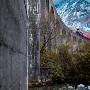 Puede incluir: Un tren rojo viaja sobre un puente de arco de piedra en un paisaje montañoso. El puente está hecho de hormigón y tiene múltiples arcos. El tren es parcialmente visible, con humo saliendo del motor. La montaña en el fondo está cubierta de nieve y niebla.