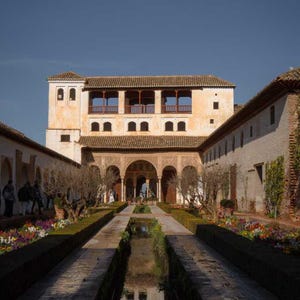May include: A view of a courtyard with a long, narrow reflecting pool in the centre. The pool is surrounded by lush greenery and stone walkways. A large, white building with arched doorways and a tiled roof is visible in the background.
