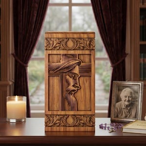 May include: A wooden urn with a carved image of Jesus on the cross. The urn is rectangular with ornate floral carvings at the top and bottom. A lit candle, a framed photo, and a book are on the table. The text on the plaque reads: Dedicated to a Life of Faith and Love.