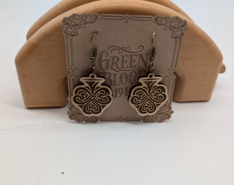 May include: Wooden earrings with a clover design. The earrings are light brown with a darker brown clover pattern. The earrings are hanging in front of a wooden display stand. The text "Green Bloom 191" is visible on the display card.