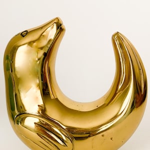 May include: A polished brass seal sculpture in a curved pose. The seal's head is raised, and its body forms a crescent shape. The sculpture has a smooth, reflective surface, with details of the seal's flippers and face.
