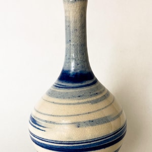 Puede incluir: Un jarrón de cerámica con base bulbosa y cuello esbelto. El jarrón presenta un cuerpo color crema con rayas azules horizontales y un cuello moteado azul y blanco. El jarrón mide aproximadamente 25 cm de altura.