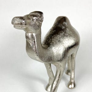 Puede incluir: Figurilla de camello plateado con una superficie detallada y texturizada. La escultura decorativa se apoya sobre cuatro patas, con la cabeza y el cuello extendidos. El camello es un objeto decorativo.