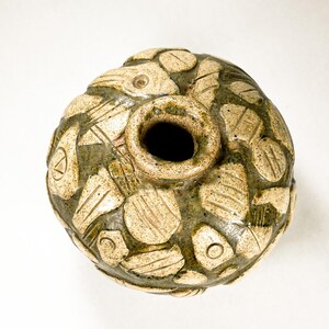 May include: A round, green and tan ceramic vase with a textured surface and a small opening at the top. The vase is decorated with a pattern of raised, geometric shapes.