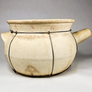 May include: A cream-colored, round ceramic cooking pot with a handle and a wire frame. The pot has a wide opening and a slightly textured surface. The wire frame appears to be made of dark metal.
