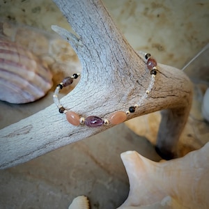 Könnte beinhalten: Ein Armband mit Perlen aus Pfirsich-, Lila- und braunen Edelsteinen. Das Armband ist an einer silbernen Kette aufgereiht und liegt auf Treibholz. Im Hintergrund sind Muscheln zu sehen. Das Armband hat eine Länge von ca. 17 cm.