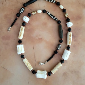 May include: A necklace featuring a mix of cylindrical bone beads with etched tan patterns, white carved bone spacers, and small black porous lava beads. The strand is accented with long, smooth dark brown beads and finished with a silver-toned metal clasp.