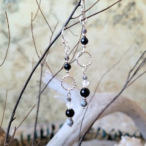 Peut inclure: Boucles d'oreilles pendantes avec crochets argentés et anneaux texturés. Chaque boucle d'oreille présente des perles noires, grises et transparentes. Les boucles d'oreilles sont présentées sur un fond flou avec des branches.