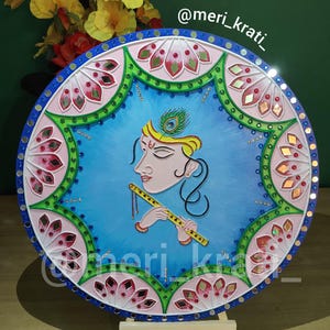 May include: A round, hand-painted artwork featuring a figure playing a flute, set against a blue backdrop. The artwork is framed with pink and green floral patterns, accented with small mirrors. The piece is displayed on a wooden easel.