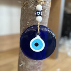 Könnte beinhalten: Ein blauer Glas-Amulett in Form eines bösen Auges mit einem weißen und blauen Augendesign, das an einer Naturfaserschnur hängt. Der Anhänger ist rund und hat eine glänzende Oberfläche. Zwei weiße Perlen und eine blaue Perle mit Augendesign befinden sich darüber.