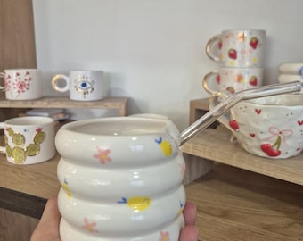 May include: A white ceramic mug with a unique, stacked design, adorned with yellow lemons and pink flowers. A clear glass straw extends from the mug. Other mugs with gold accents and fruit designs are visible on wooden shelves.