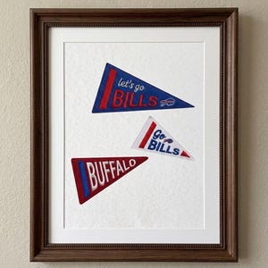 May include: A dark brown wooden frame containing three embroidered pennants on a white background. One blue pennant reads "let's go BILLS", one white pennant reads "Go BILLS", and one red pennant reads "BUFFALO". Each pennant features team logos.