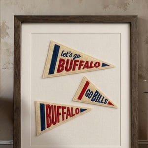 Puede incluir: Una pieza de arte mural enmarcada que presenta tres banderines de fieltro color crema con bordados en rojo y azul. Los banderines muestran el texto 'let's go BUFFALO', 'GO BILLS' y 'BUFFALO' en una disposición triangular en un marco de madera oscura.