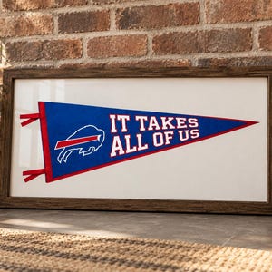 Pode incluir: Uma peça de arte de parede retangular emoldurada com um galhardete triangular azul com acabamento vermelho. O galhardete exibe um logótipo branco dos Buffalo Bills e o texto IT TAKES ALL OF US em letras maiúsculas brancas. A moldura é de madeira castanha escura.