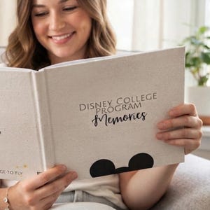 May include: A light beige hardcover book with the text "DISNEY COLLEGE PROGRAM Memories" printed in black on the front cover. The cover features a black silhouette of mouse ears at the bottom and the text "GE TO FLY" in the corner.