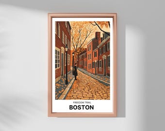 May include: Framed art print featuring a street scene from Boston's Freedom Trail. The artwork depicts brick buildings, a cobblestone street, and a person standing near a lamppost. The print has a rose-gold frame and the text "FREEDOM TRAIL BOSTON".