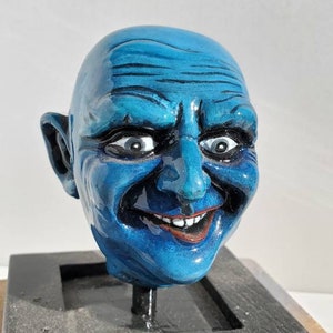 May include: A blue, smiling face sculpture with a bald head and detailed facial features. The sculpture has a glossy finish and is mounted on a black, rectangular base. The eyes are wide and the mouth is open, revealing red lips.